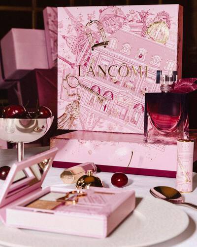 Празнична аранжировка на празничната колекция на Lancome върху маса, с голяма илюстрована розова подаръчна кутия, парфюм, грим продукти и свежи череши.