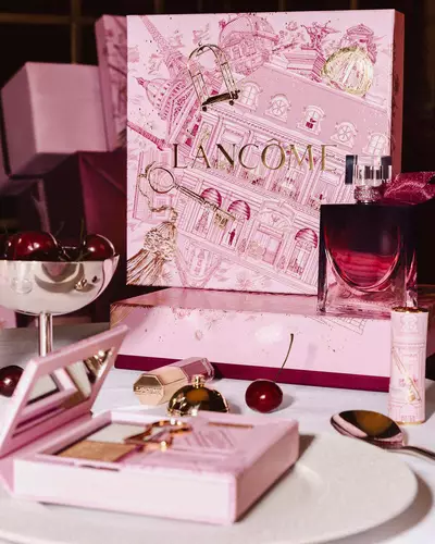 Празнична аранжировка на празничната колекция на Lancome върху маса, с голяма илюстрована розова подаръчна кутия, парфюм, грим продукти и свежи череши.