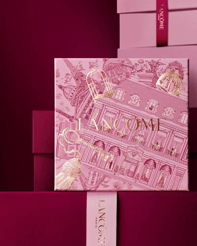 Изображение на розова празнична подаръчна кутия Lancome пред редица от розови и тъмночервени подаръчни кутии, разположени на тъмночервен фон.