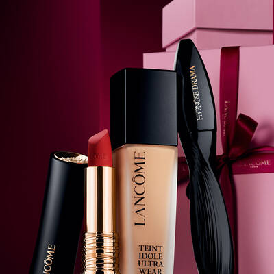 Елегантен натюрморт от матово червило L'Absolu Rouge на Lancome, фон дьо тен Teint Idole Ultra Wear и спирала Hypnose Drama, поставени върху луксозен тъмночервен празничен фон.