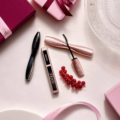 Подредба отгоре на грим продукти Lancome, включваща спирала и гланц за устни, заедно с розови подаръчни кутии, сатенени панделки и стръкче френско грозде.
