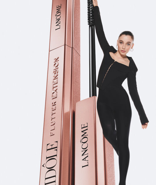 Натурална жена позира с новата спирала за мигли на Lancome на фона на стилен бял фон.