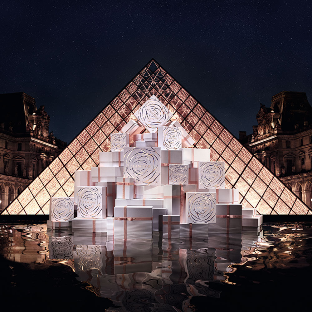 A pyramid of beautifully wrapped gift boxes with rose designs is displayed in front of the illuminated glass pyramid of the Louvre at night, reflecting in the water below.