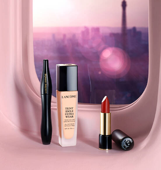 A vanity display of Lancome makeup products, featuring a liquid foundation, a black eyeliner, and a red lipstick, with a blurred cityscape in the background.