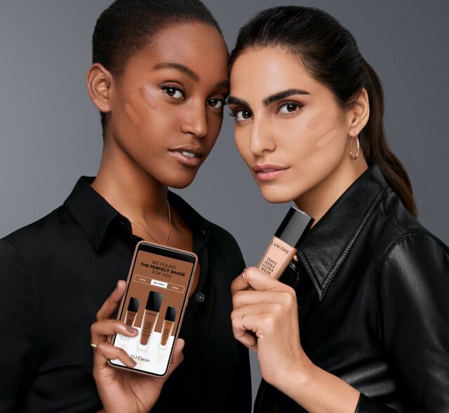 Two women of different ethnicities hold makeup products, standing against a dark gray background. One woman is displaying a smartphone with foundation shades, while the other holds a tube of foundation.