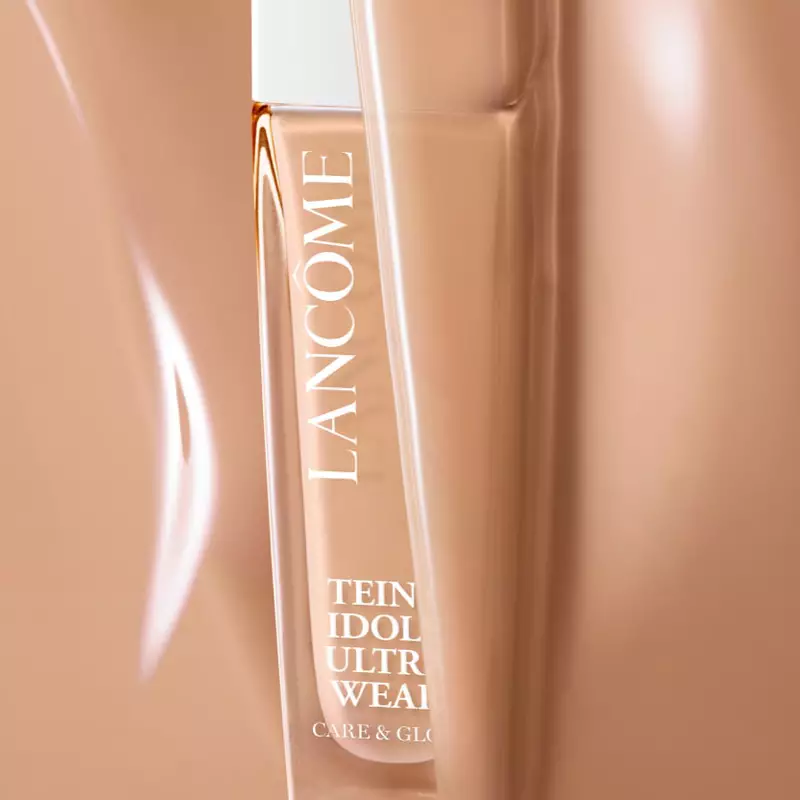 A close-up of a Lancome foundation bottle partially obscured in a warm-toned background. The bottle is sleek and elegant, labeled "Teint Idole Ultra Wear."