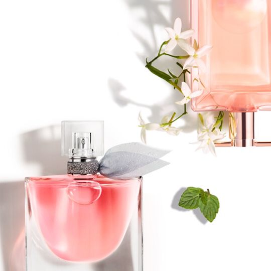 A close-up of two elegant perfume bottles surrounded by white flowers and a green mint leaf, set against a white background.