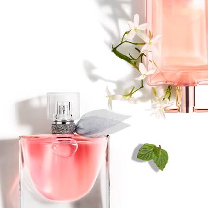A close-up of two elegant perfume bottles surrounded by white flowers and a green mint leaf, set against a white background.