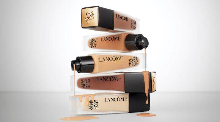 A stack of Lancome foundation bottles in various shades, with some bottles tilted and liquid foundation dripping from them, set against a neutral background.