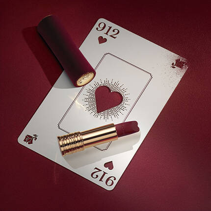 A playing card with a heart design lies on a deep red surface, next to a tube of lipstick in a matching shade. The card features ornate details, enhancing the elegant aesthetic.