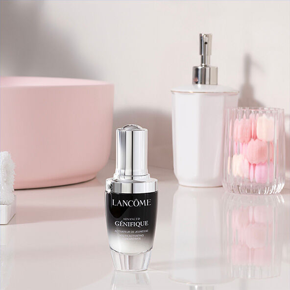 A bottle of Lancome Advanced Genifique serum is placed on a glossy surface, surrounded by soft pink decor and a small vase with a blooming rose.