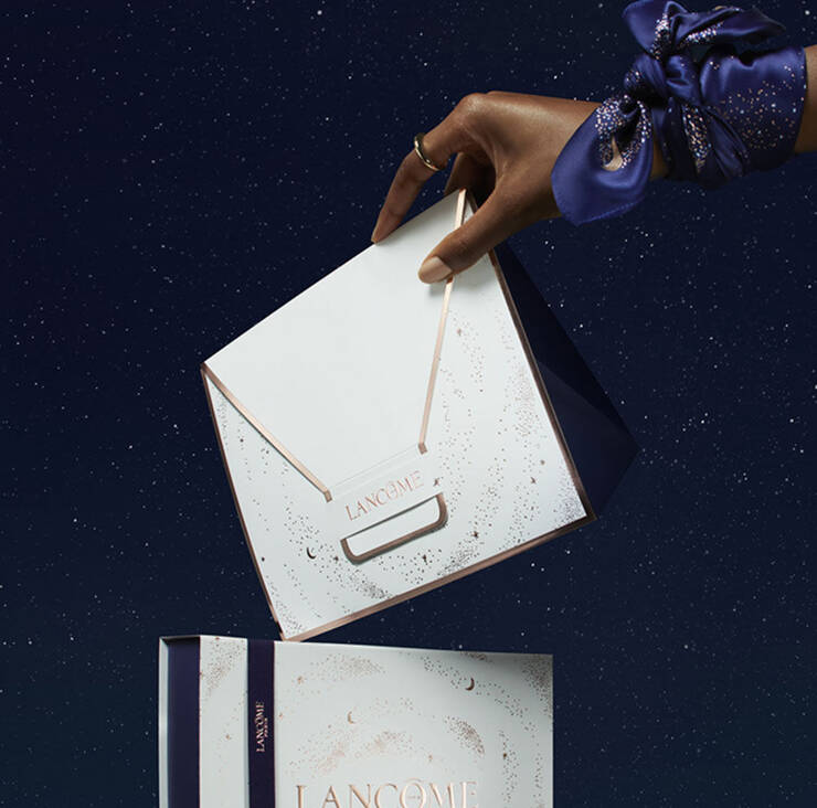 A hand elegantly holds a white envelope with gold accents labeled "Lancome," above a beautifully designed gift box set against a starry night background.