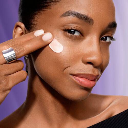 A woman applies skincare cream to her cheek, smiling confidently, with a blurred purple background.