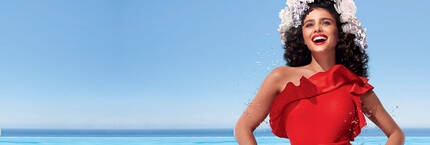 A woman with curly hair, wearing a red dress and a flower crown, smiles joyfully against a bright blue sky and ocean backdrop.