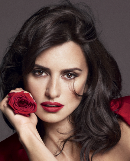 A woman with dark hair and red lipstick holds a red rose while wearing a bold red outfit against a neutral background.