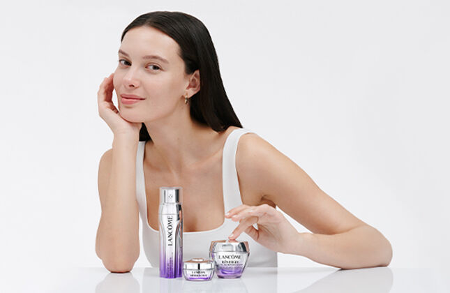 A young woman with long dark hair sits at a white table, smiling softly as she showcases skincare products in front of her. The background is light and minimalistic.