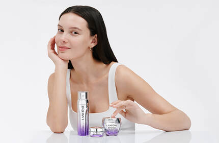 A young woman with long dark hair sits at a white table, smiling softly as she showcases skincare products in front of her. The background is light and minimalistic.
