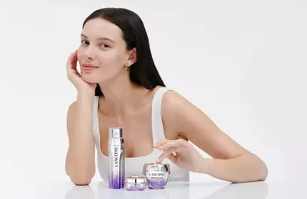 A young woman with long dark hair sits at a white table, smiling softly as she showcases skincare products in front of her. The background is light and minimalistic.