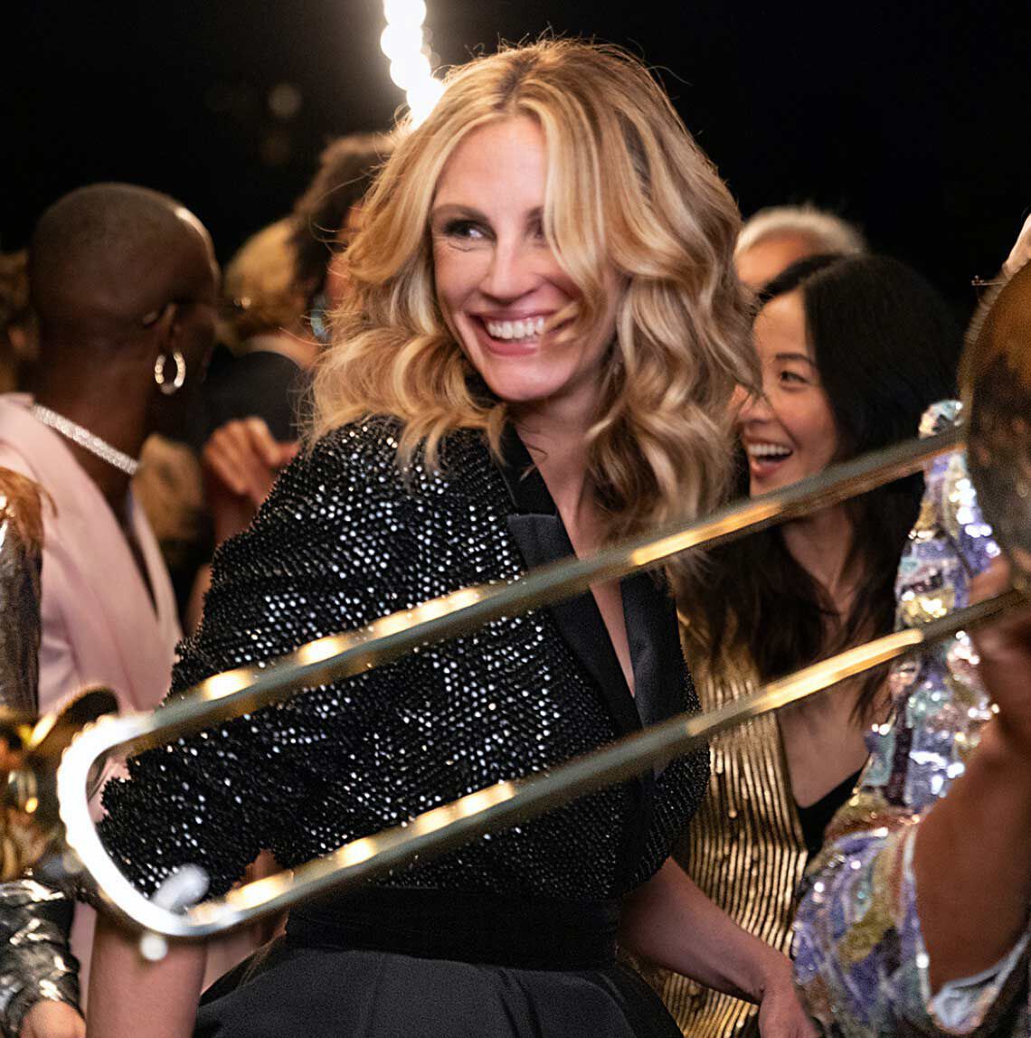 Упоследна сцена на парти с усмихната жена с вълнисти коси, облечена в бляскав черен сако, заобиколена от други хора, които се забавляват.