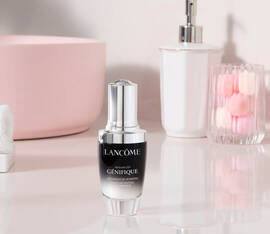 A bottle of Lancome Advanced Genifique serum is placed on a countertop beside a glass vase with a pink rose and decorative balls, with a light pink bowl and a rolled towel in the background.