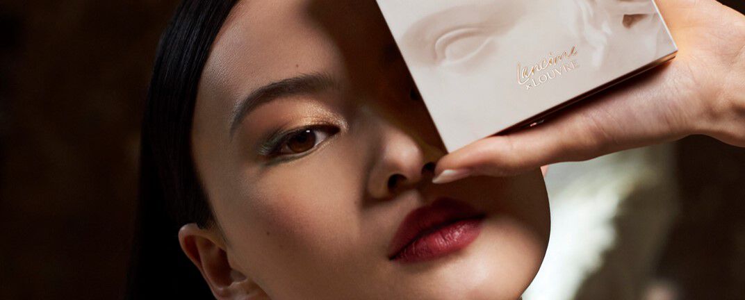 A close-up of a model's face, holding a beauty product close to her cheek. She has a bold lip color and subtle eye makeup, with a soft expression. The background is softly blurred.