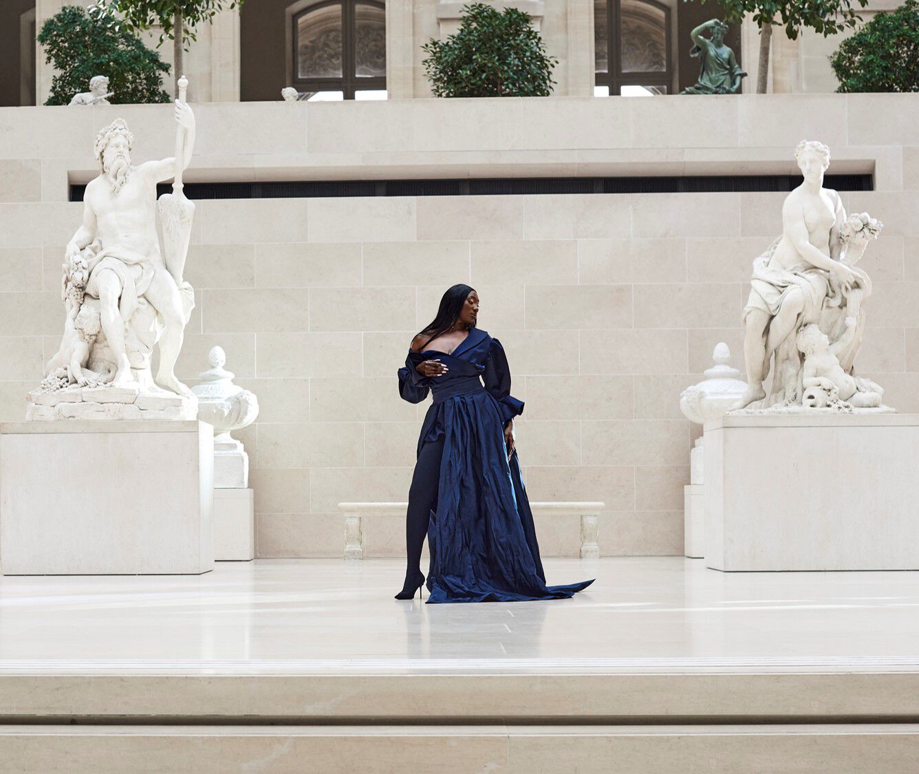 A model in a flowing navy gown stands elegantly on a marble platform surrounded by classical sculptures in a spacious, well-lit gallery.